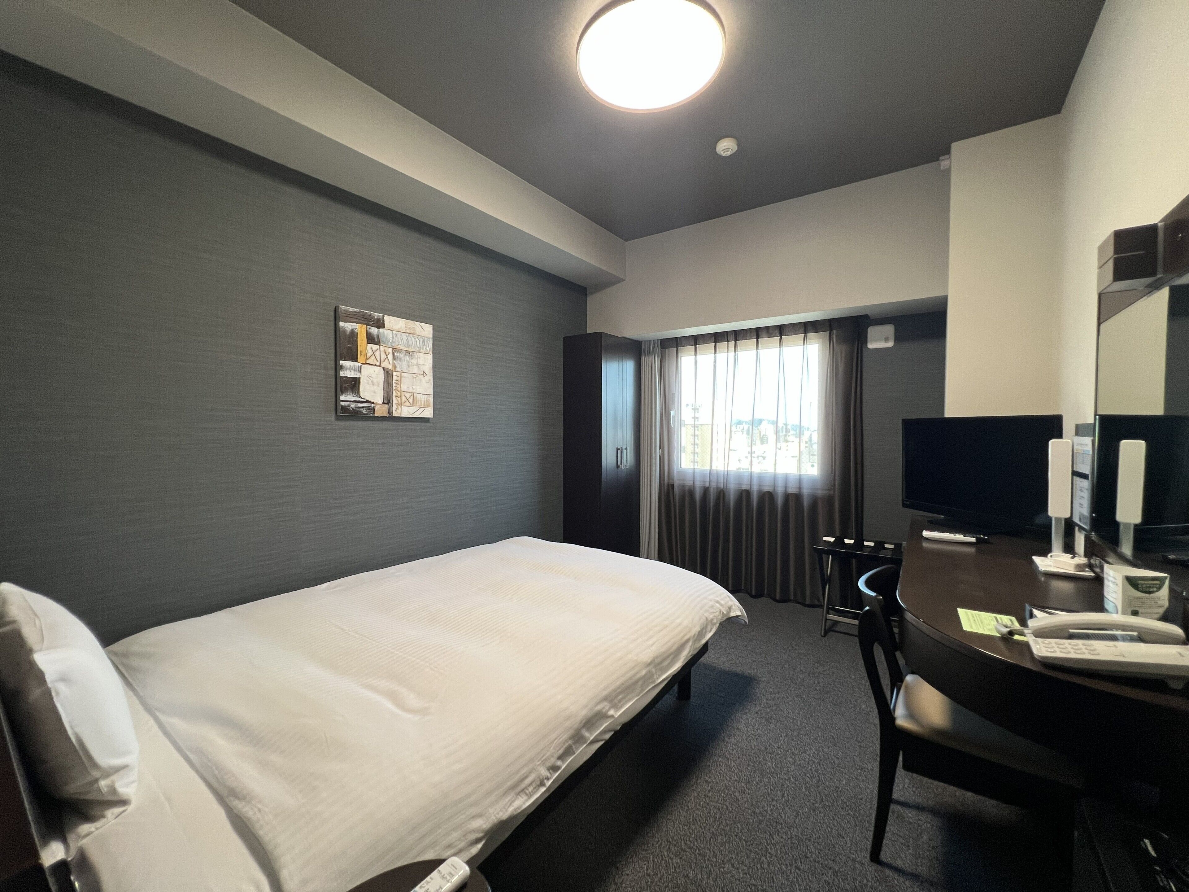 Habitación de hotel con cama, escritorio, teléfono, televisor y ventana con cortinas.