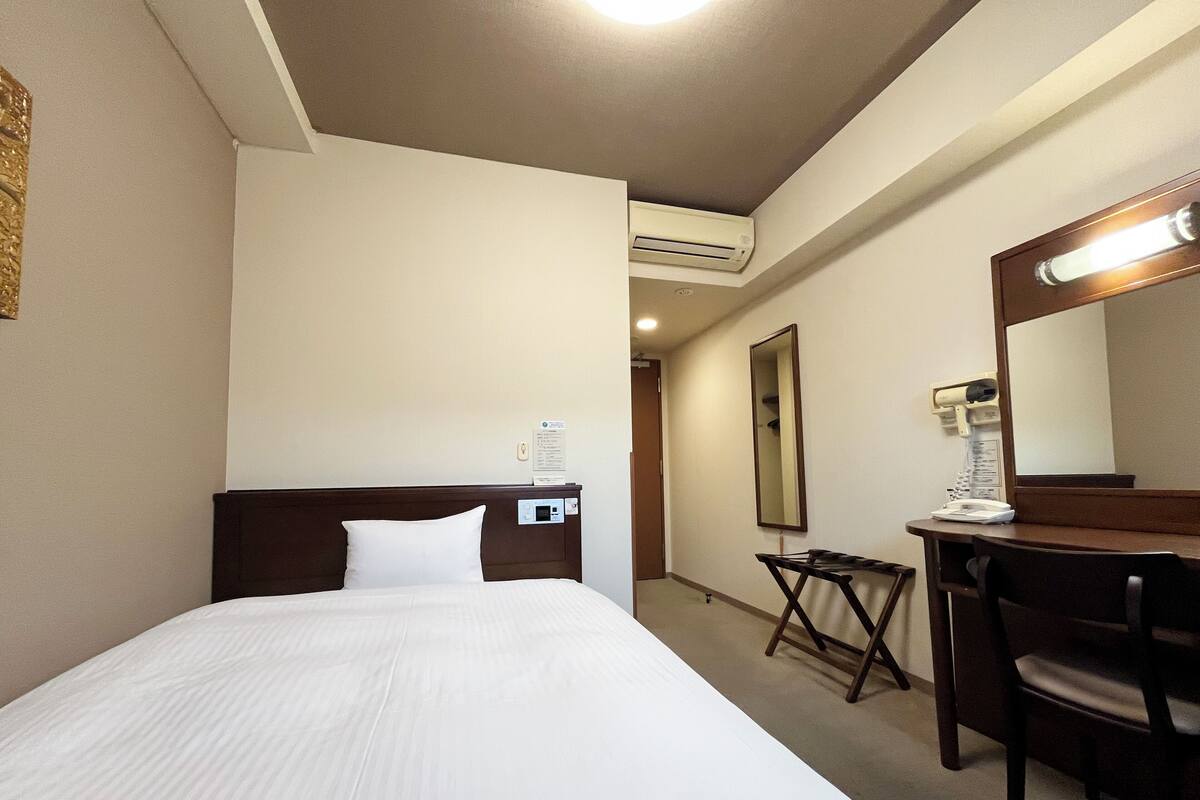 A hotel room with a bed, a desk, a chair, a mirror, and an air conditioning unit.