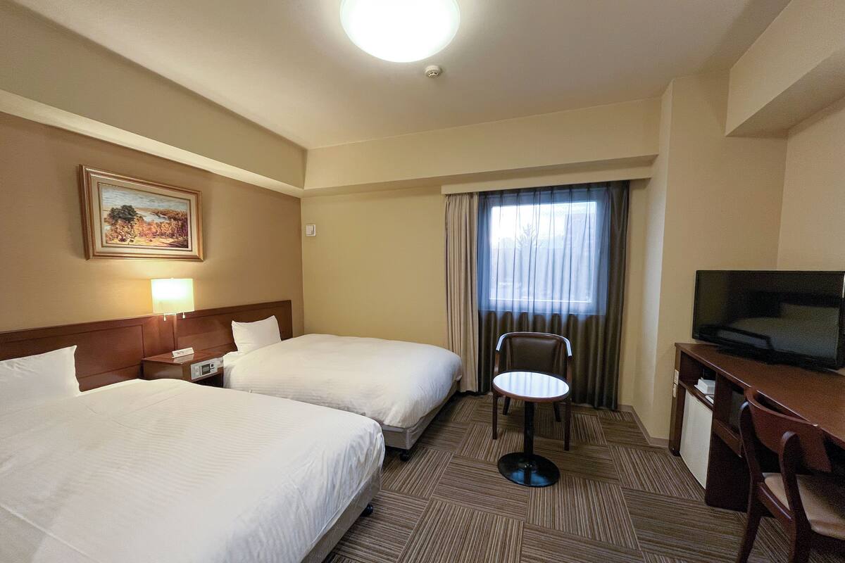A hotel room with two beds, a desk with a television, a chair, a lamp, and a painting on the wall.