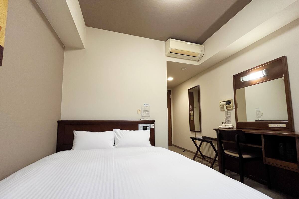 A hotel room with a bed, a desk, a mirror, and an air conditioning unit.