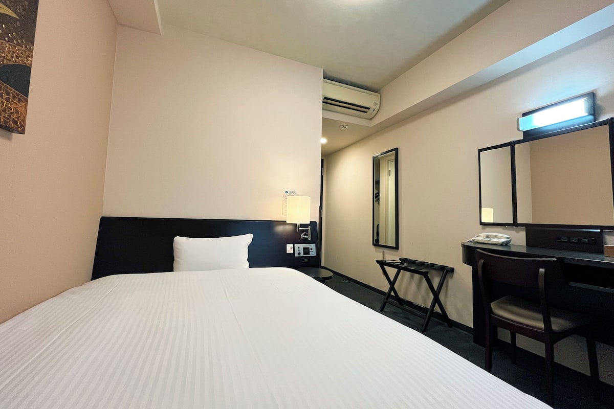 A hotel room with a large bed, a desk with a chair, a desk lamp, a telephone, and a mirror.