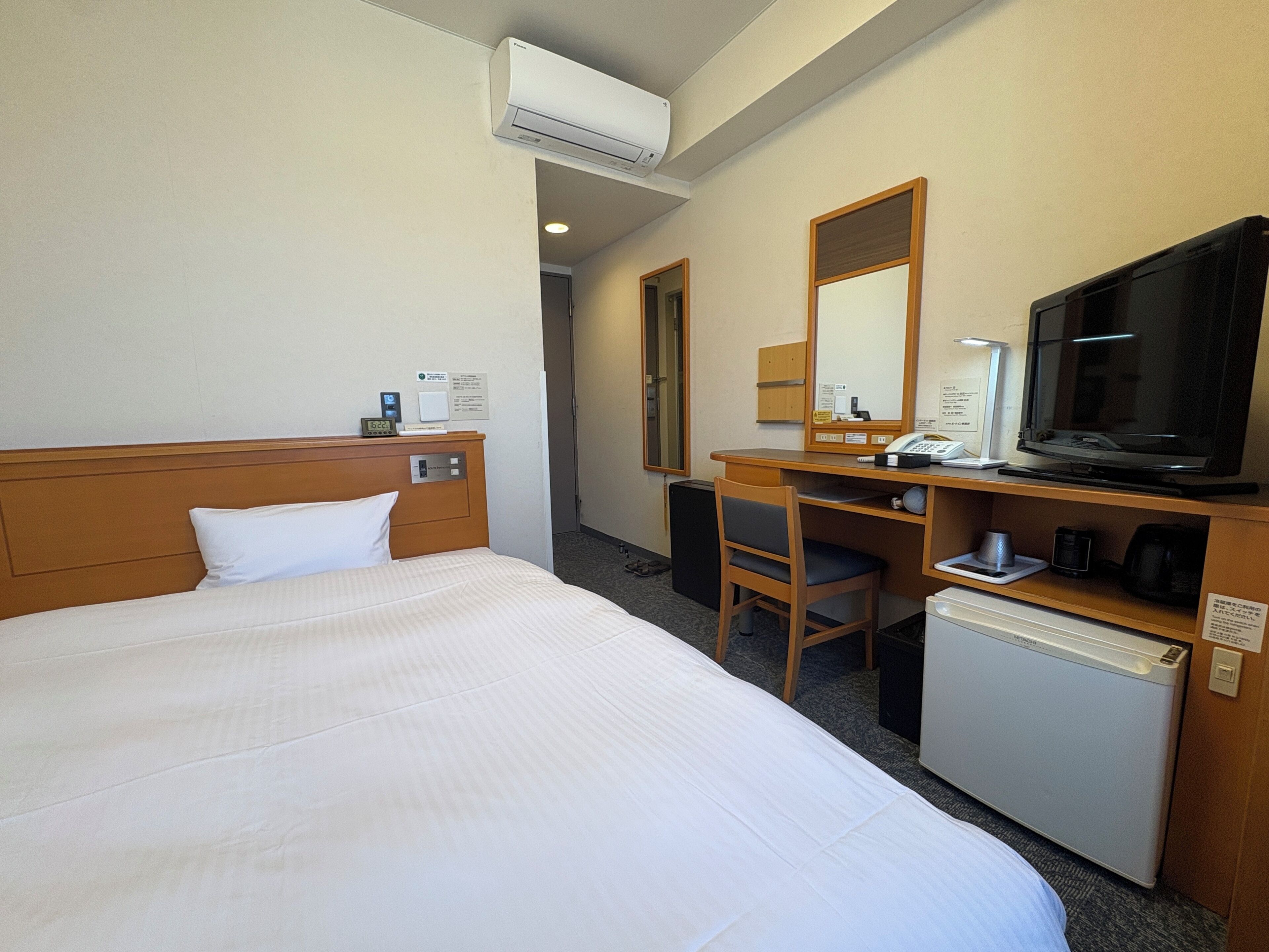 A hotel room with a bed, a desk, a television, a coffee maker, and an air conditioning unit.