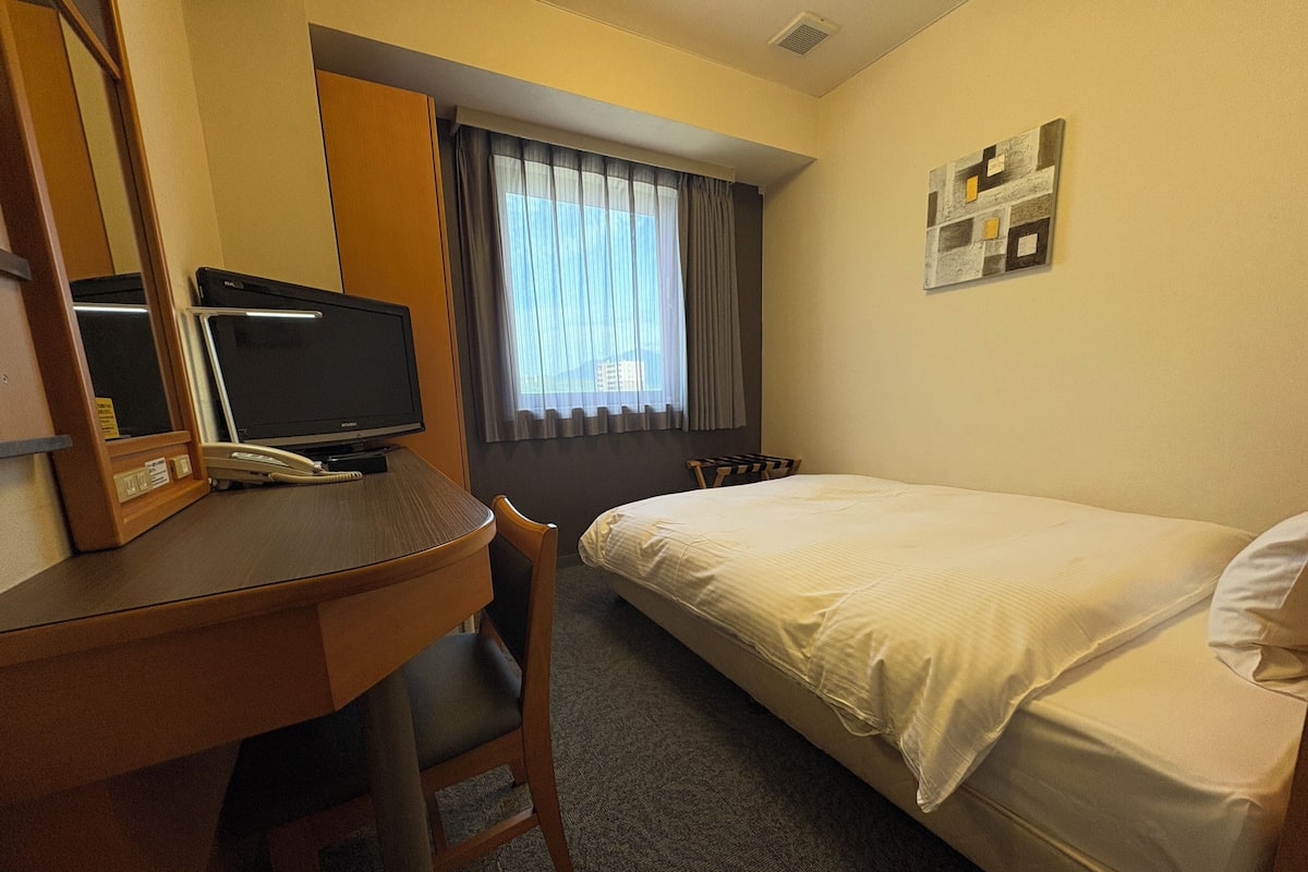 Una habitación de hotel con cama, escritorio, teléfono, televisor, silla y ventana con cortinas.