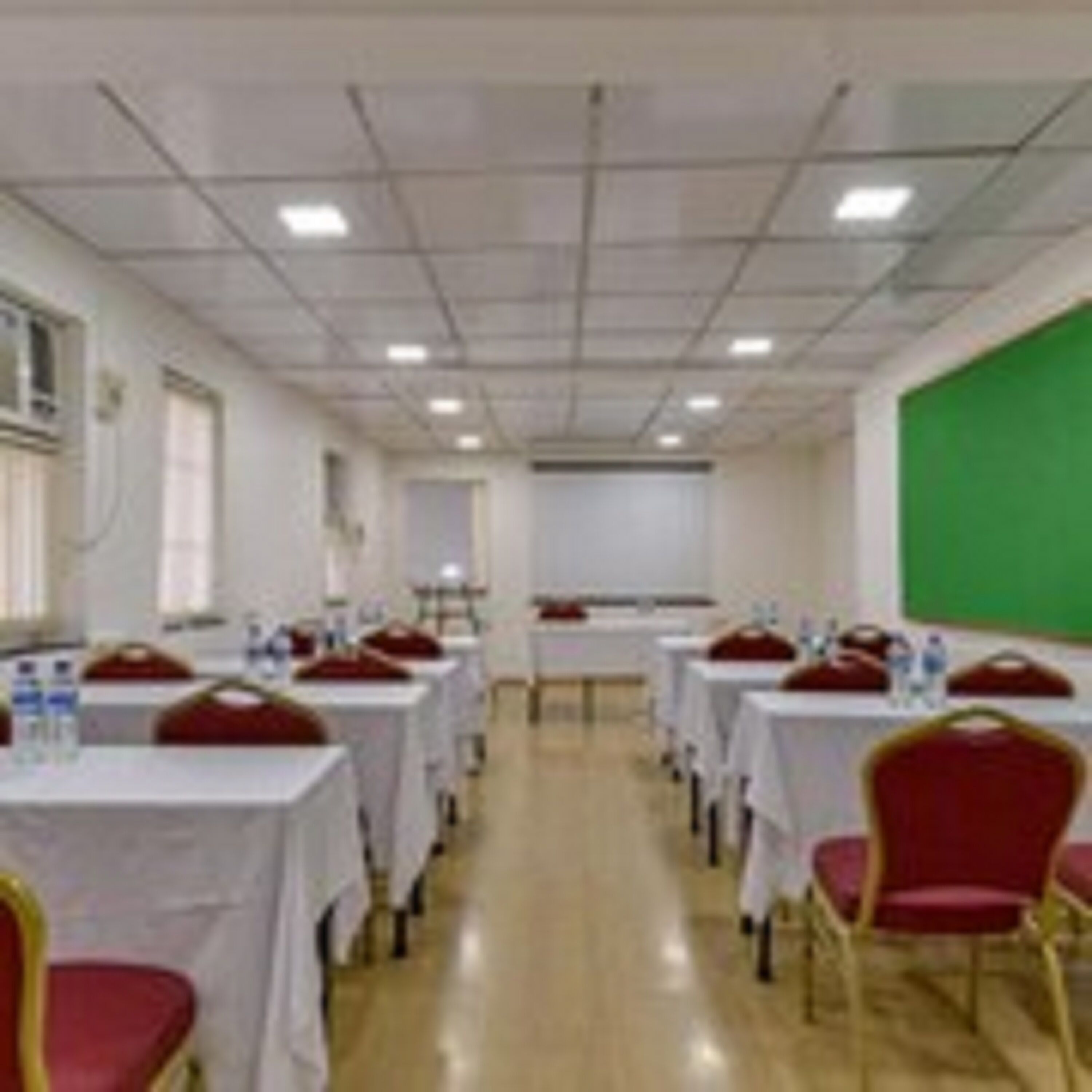 Un salón de conferencias con filas de mesas y sillas, equipado con botellas de agua y una pantalla verde.