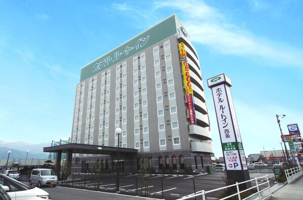 A multi-story hotel with a green sign at the top, a parking lot in front, and a tall sign with Japanese text to the right. - Hotel Route Inn Iyo Saijo (Saijo)