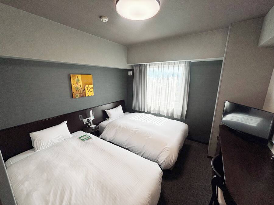 A hotel room with two beds, a bedside table with a phone and a lamp, a window with curtains, and a painting on the wall.