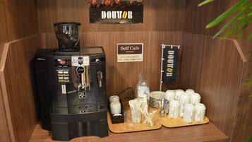 Una estación autoservicio de café con una máquina de café, tazas y un cartel con el horario de funcionamiento.
