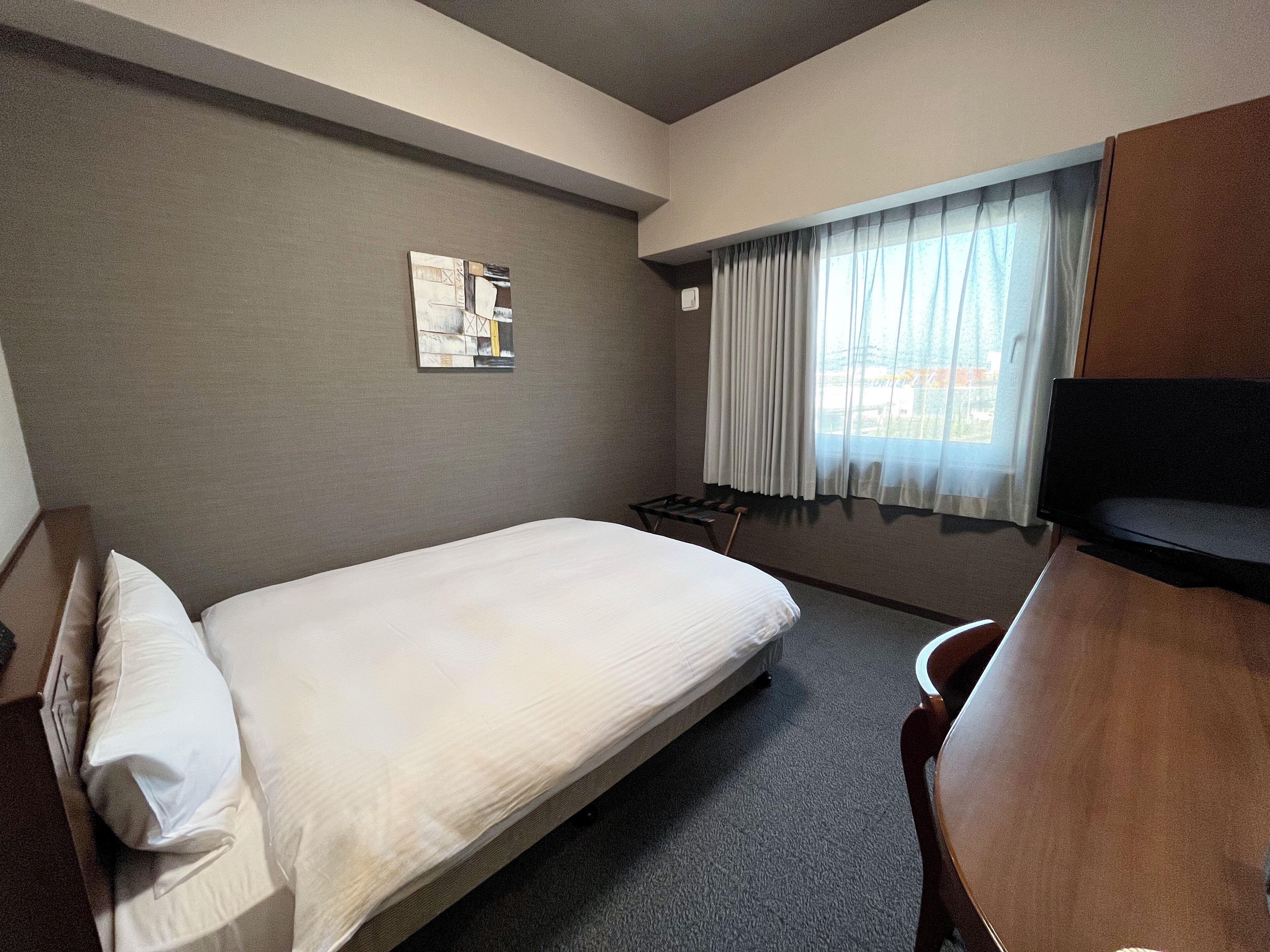 Quarto de hotel com uma cama de solteiro, escrivaninha, cadeira, janela com cortinas e uma obra de arte na parede.
