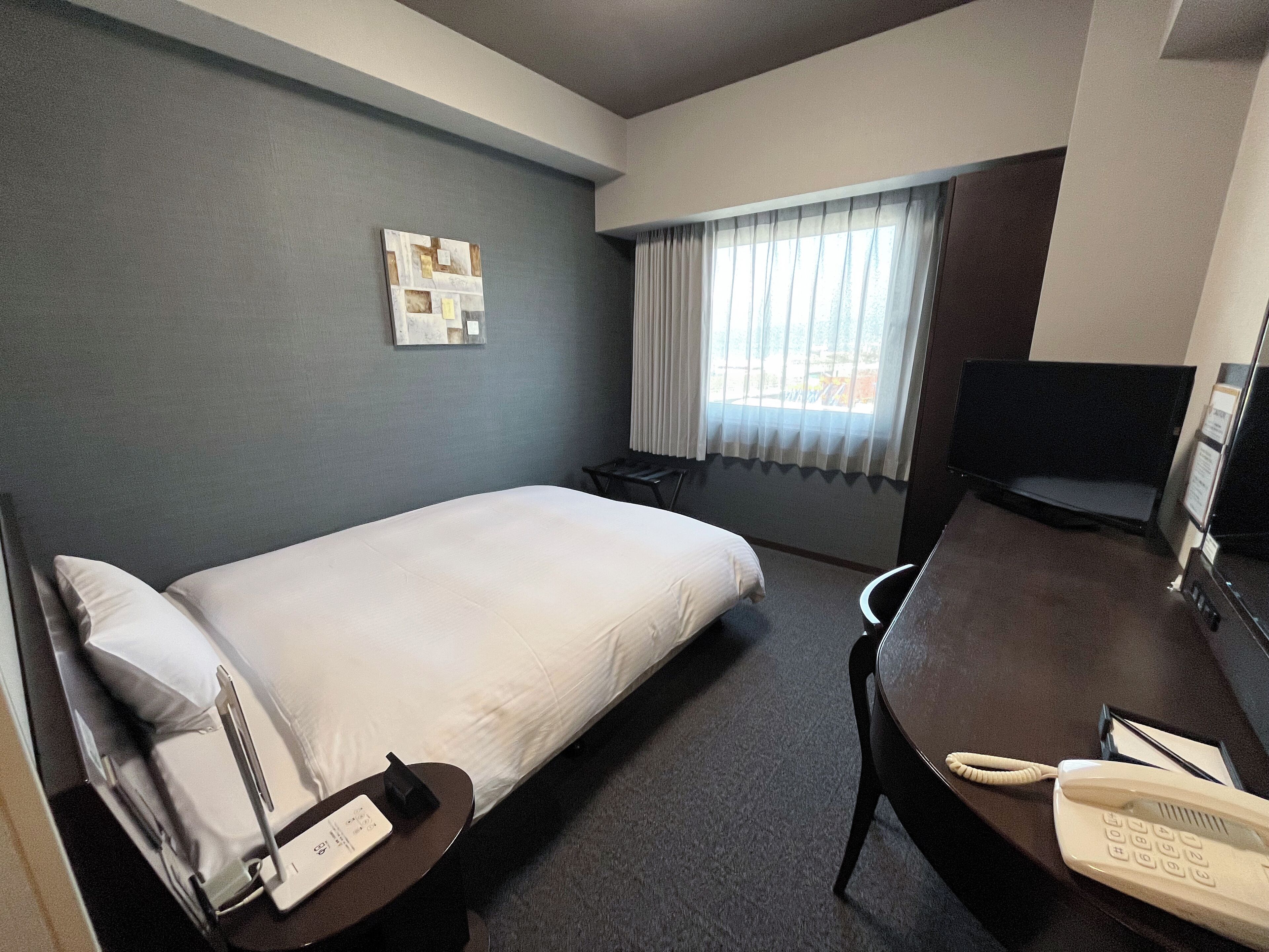 Ein Hotelzimmer mit einem Einzelbett, einem Schreibtisch mit Telefon, einem Sessel, einem kleinen Tisch, einem Fernseher und einem Fenster mit Vorhängen.