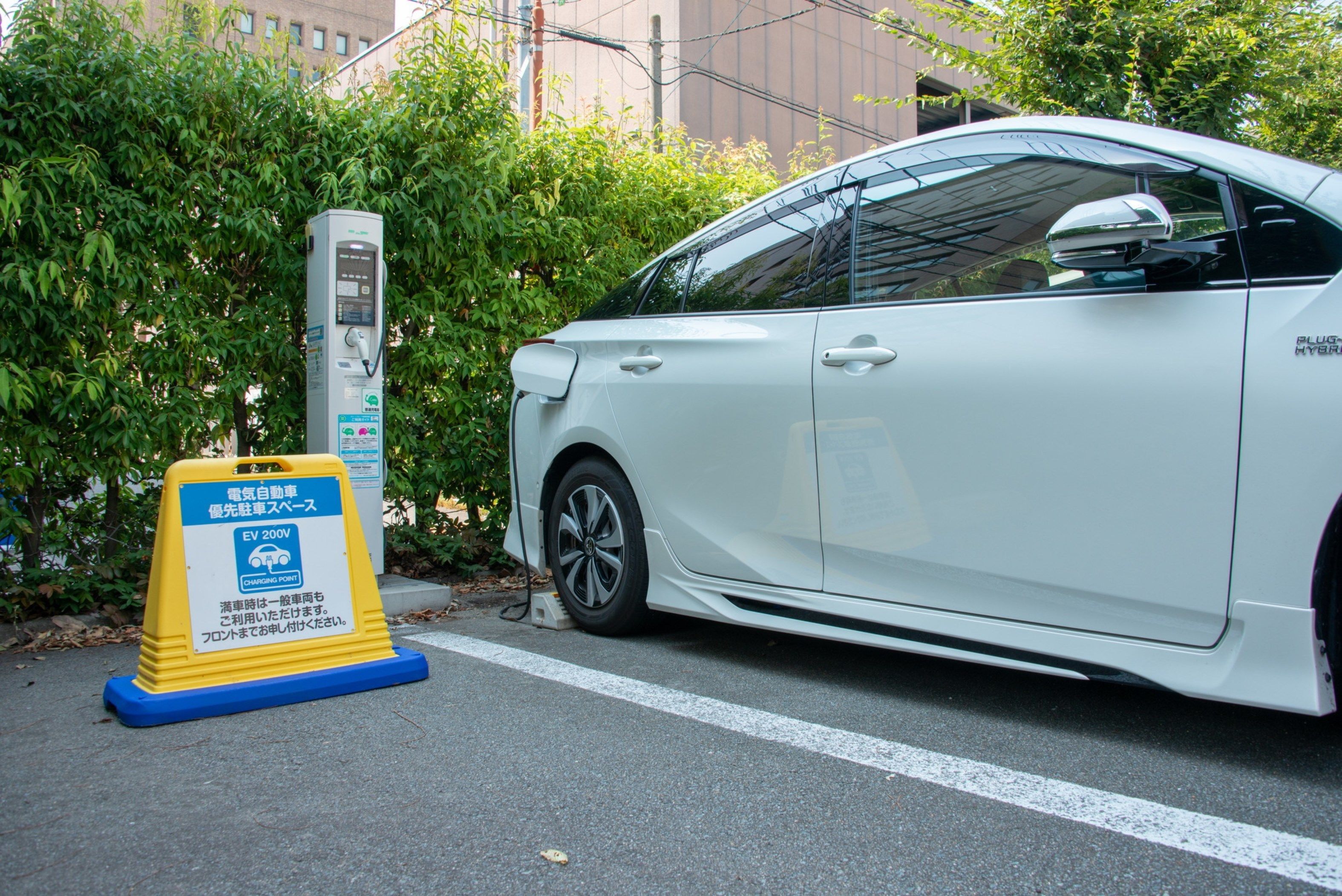 Un vehículo eléctrico blanco cargándose en una estación con un letrero amarillo que indica EV 200V.