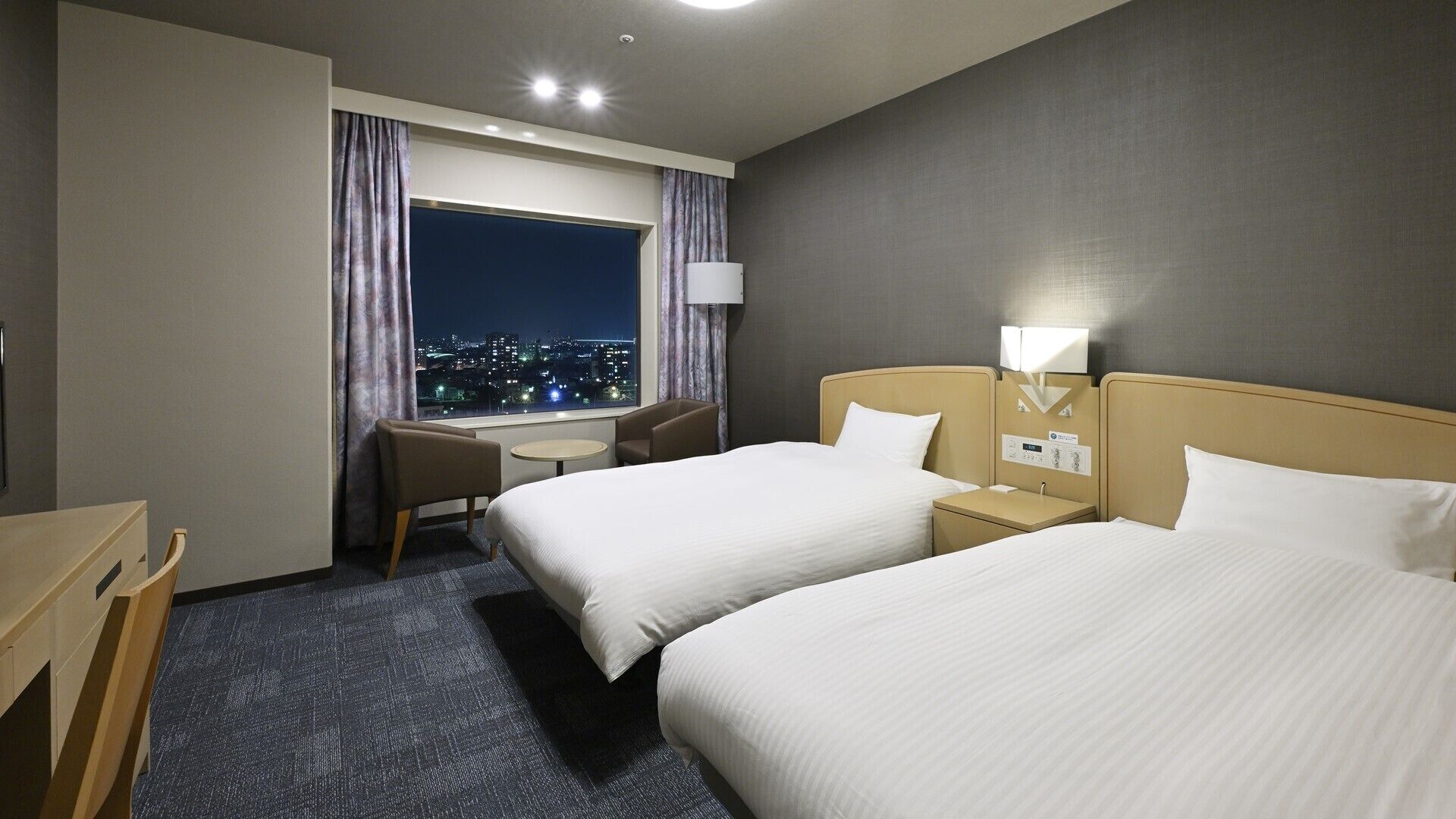 A hotel room with two beds, a desk, a chair, a window with a city view, and a night sky visible through the window.