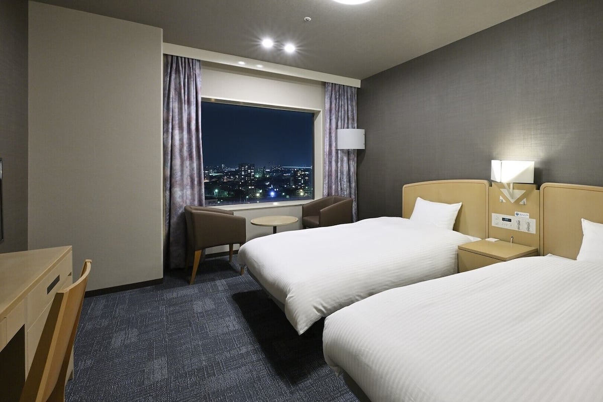 Ett hotellrum med två sängar, ett skrivbord, en stol, ett fönster med utsikt över staden och en stjärnklar natthimmel som syns genom fönstret.