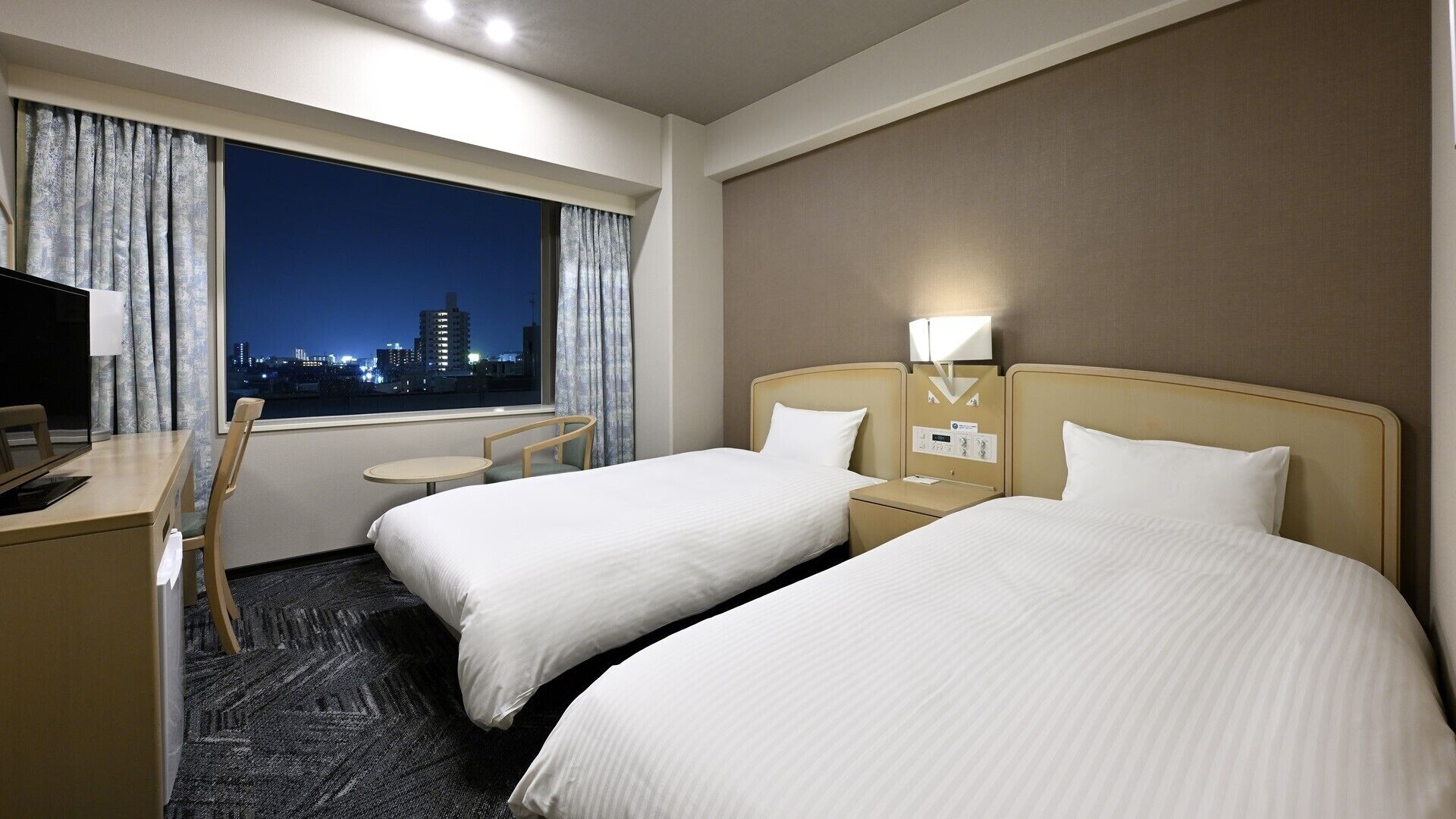 A hotel room with two beds, a desk, a chair, a small table, and a window with a city view.