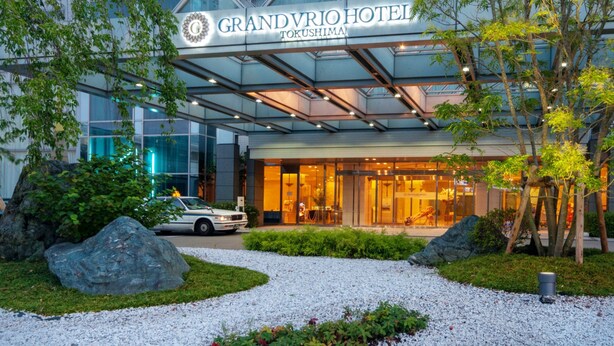 The GrandVrio Hotel Tokushima exterior with a modern glass facade, large rock landscaping, and a car parked in front.