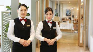 Two waitresses in a hotel lobby with a modern interior, a reception desk, and a dining area in the background.