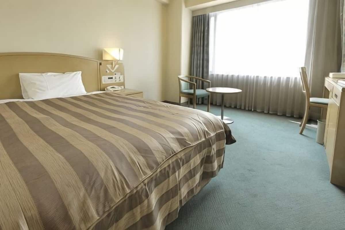 Ein Hotelzimmer mit einem groĂen Bett, einem Schreibtisch mit Stuhl, einem kleinen Tisch und einem Fenster mit VorhĂ€ngen.