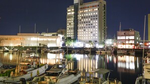 A marina with boats docked, a tall hotel building with illuminated windows, and a calm water body reflecting the lights.