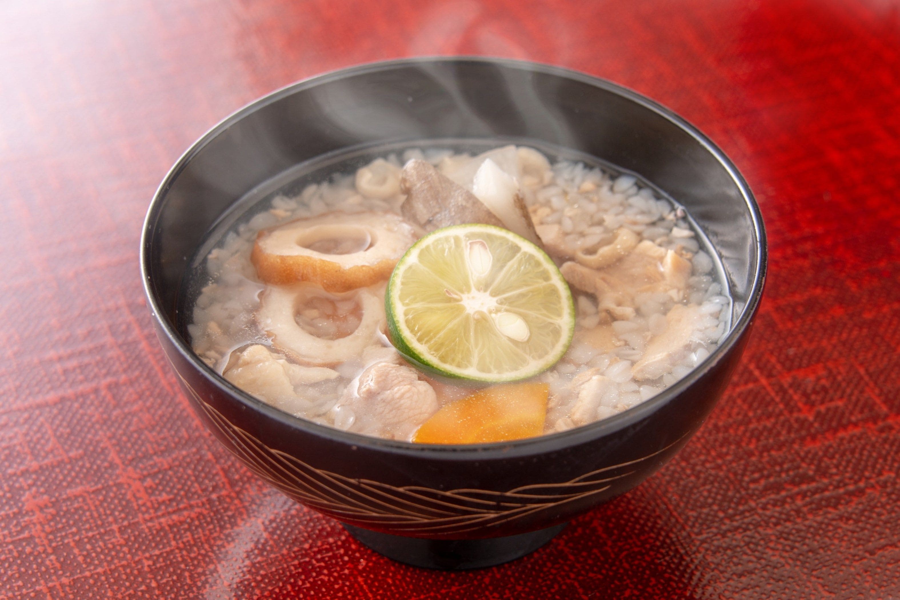 A bowl of soup with various ingredients, including meat and vegetables, on a red textured table.