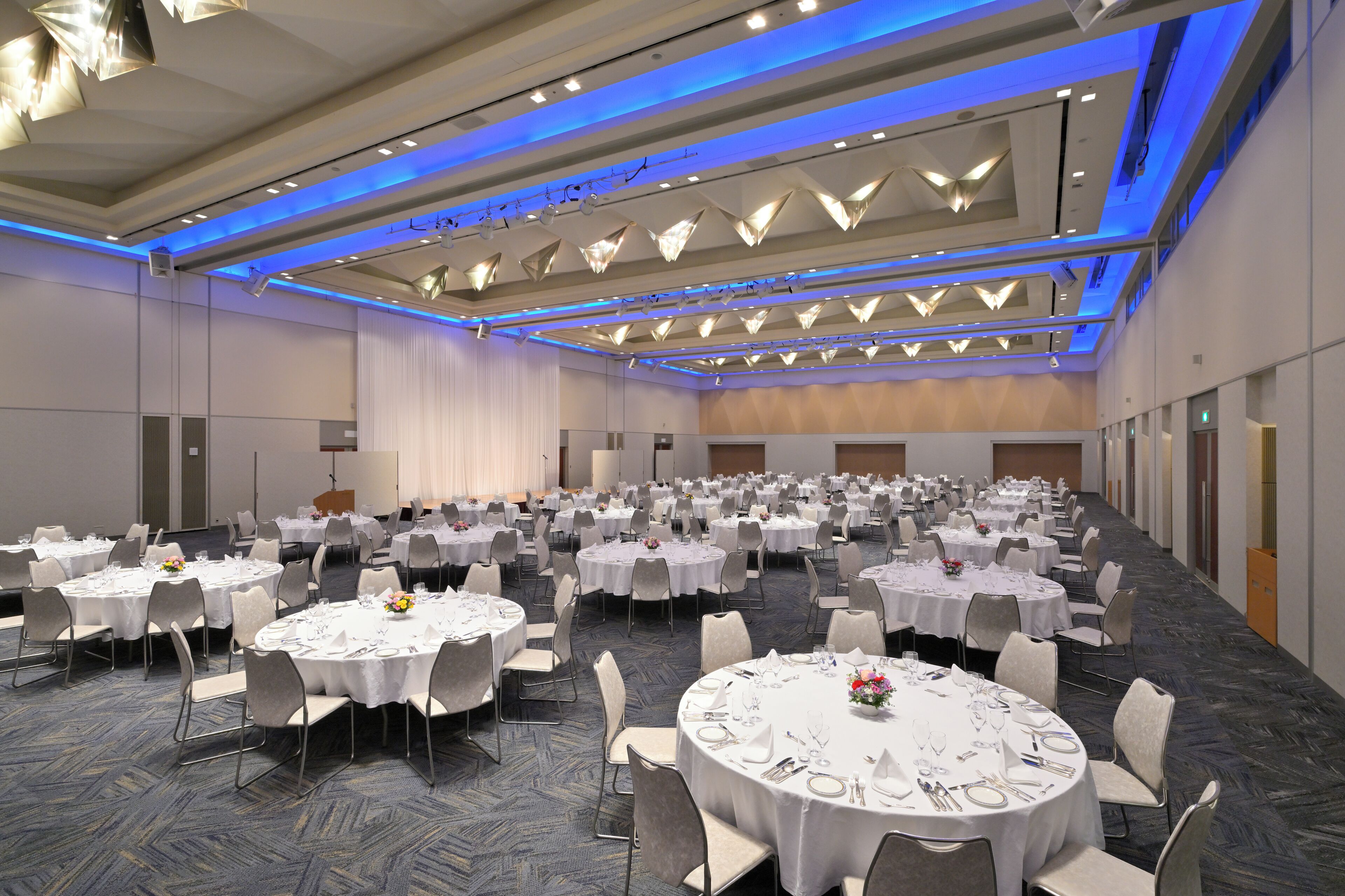 Un amplio salón de banquetes con varias mesas redondas, cada una con mantel blanco, sillas y vajilla.