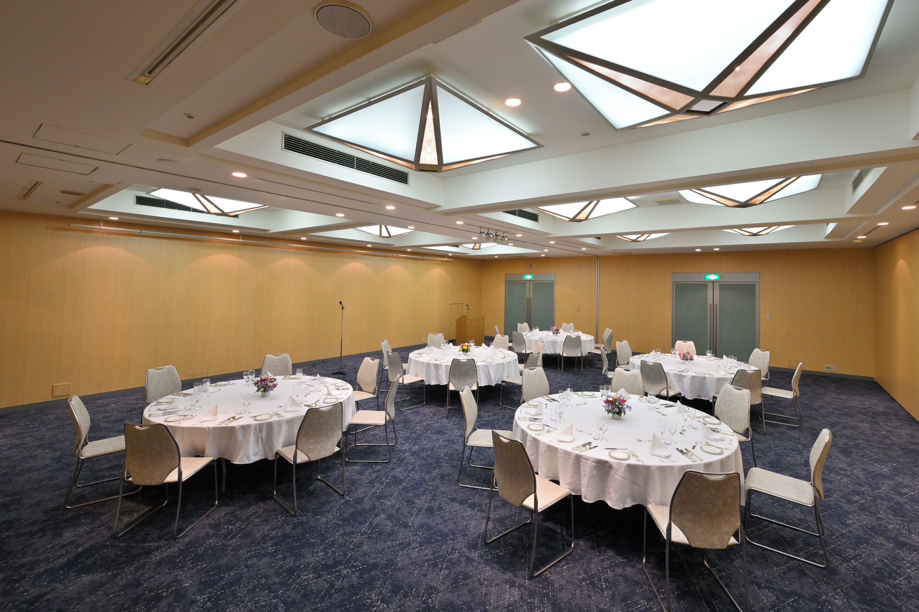 A conference room with multiple round tables, each set with white tablecloths, chairs, and centerpieces. The room features a high ceiling with skylights and recessed lighting.