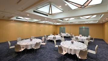 A conference room with multiple round tables, each set with white tablecloths, chairs, and centerpieces. The room features a high ceiling with skylights and recessed lighting.