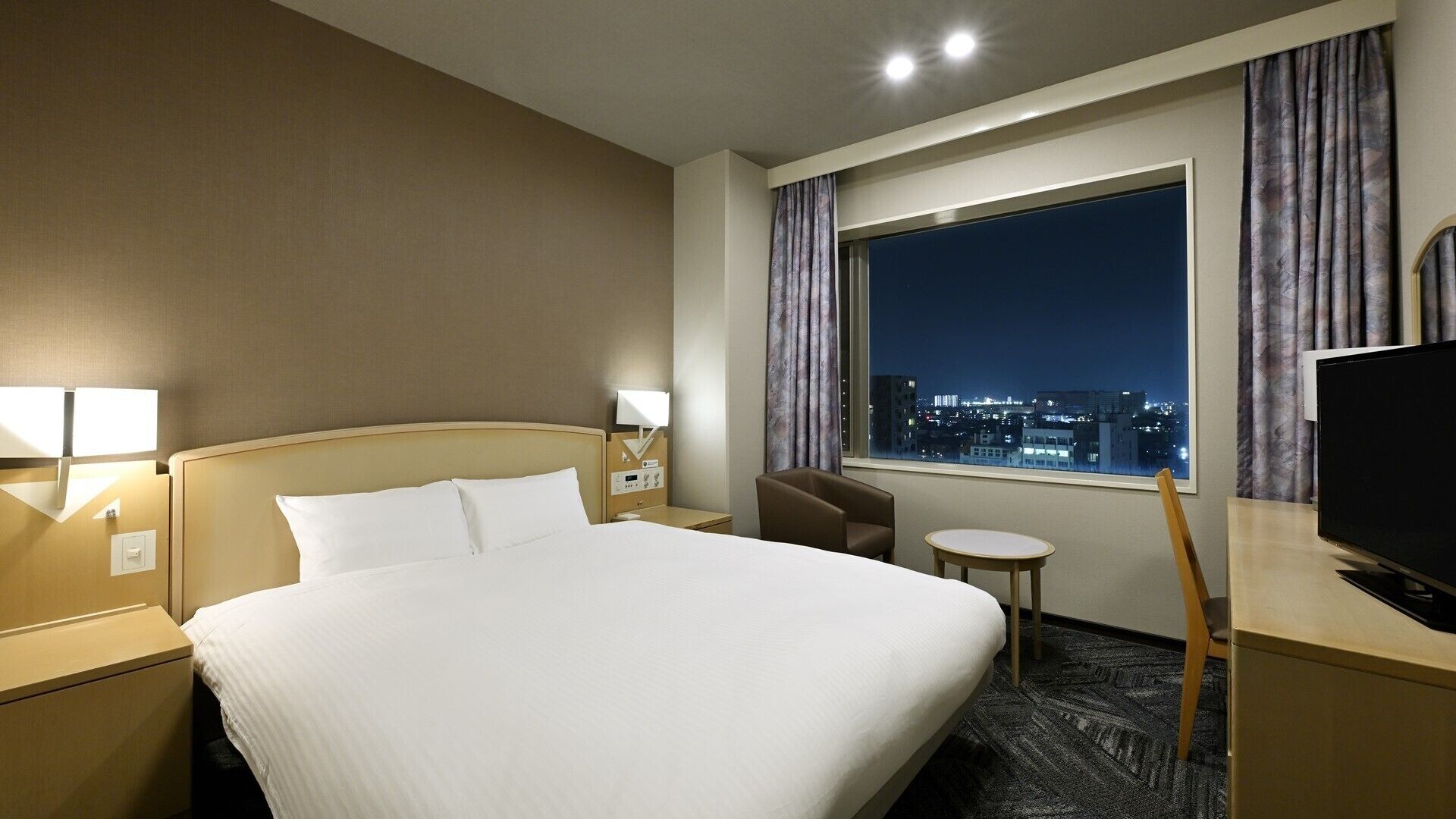 A hotel room with a bed, bedside lamps, a desk with a TV, a chair, a small table, and a window with a city view.