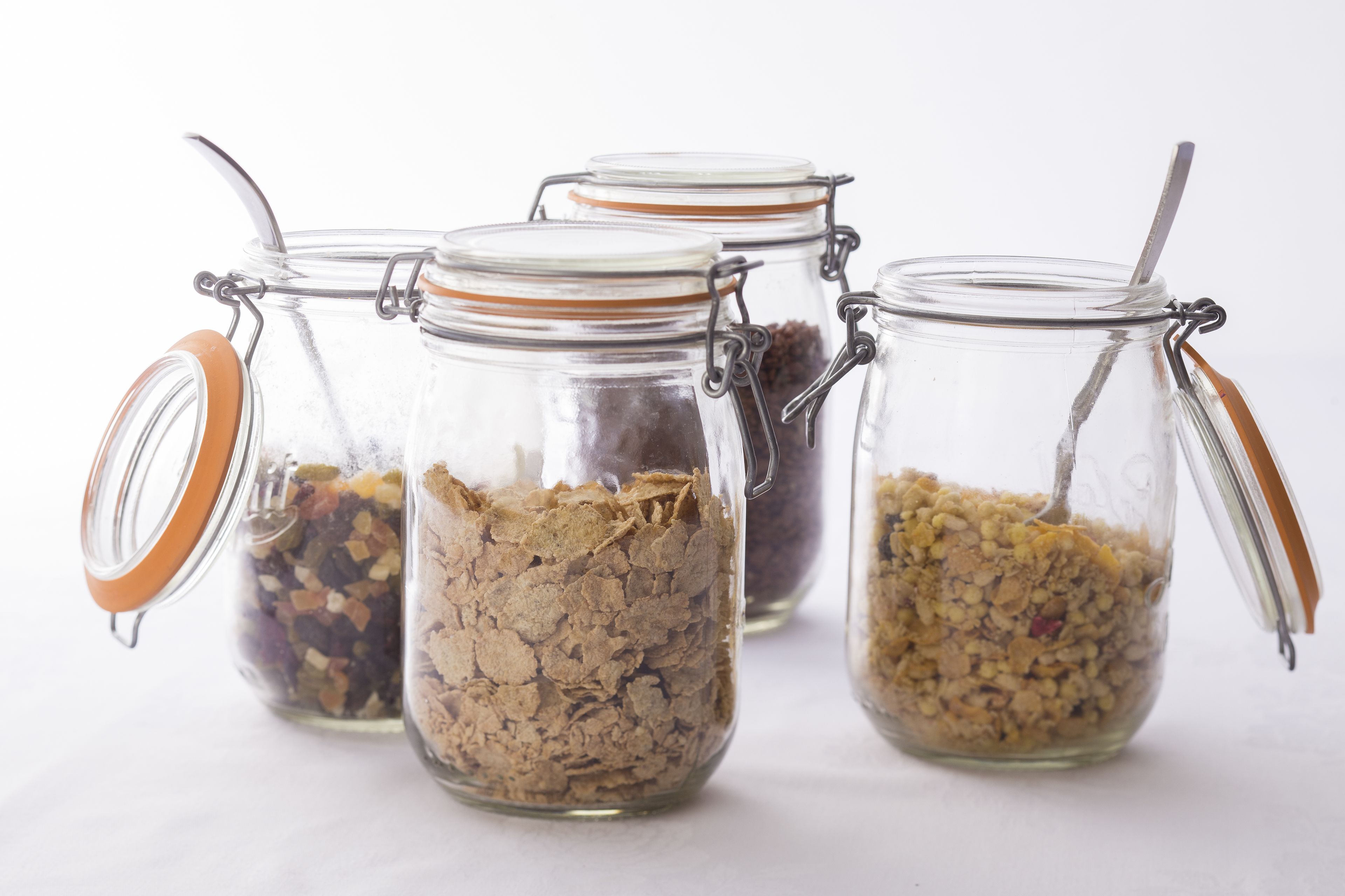 Cuatro frascos de vidrio con cierres metálicos y asas anaranjadas, que contienen diversos cereales y semillas.