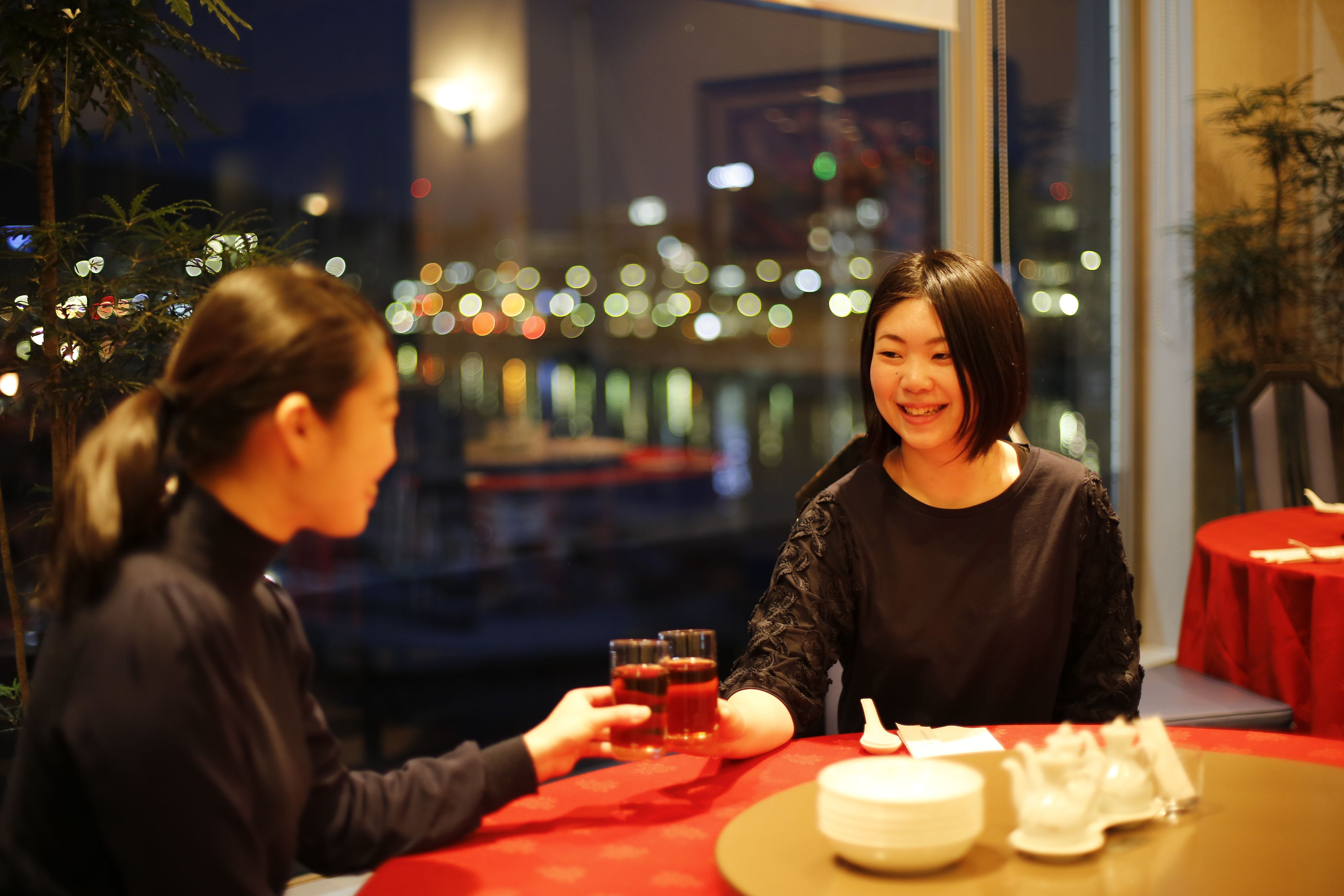 Two people seated at a table with a red tablecloth, holding glasses of red beverage. The setting includes a view of city lights through a window, and a potted plant is visible on the right.