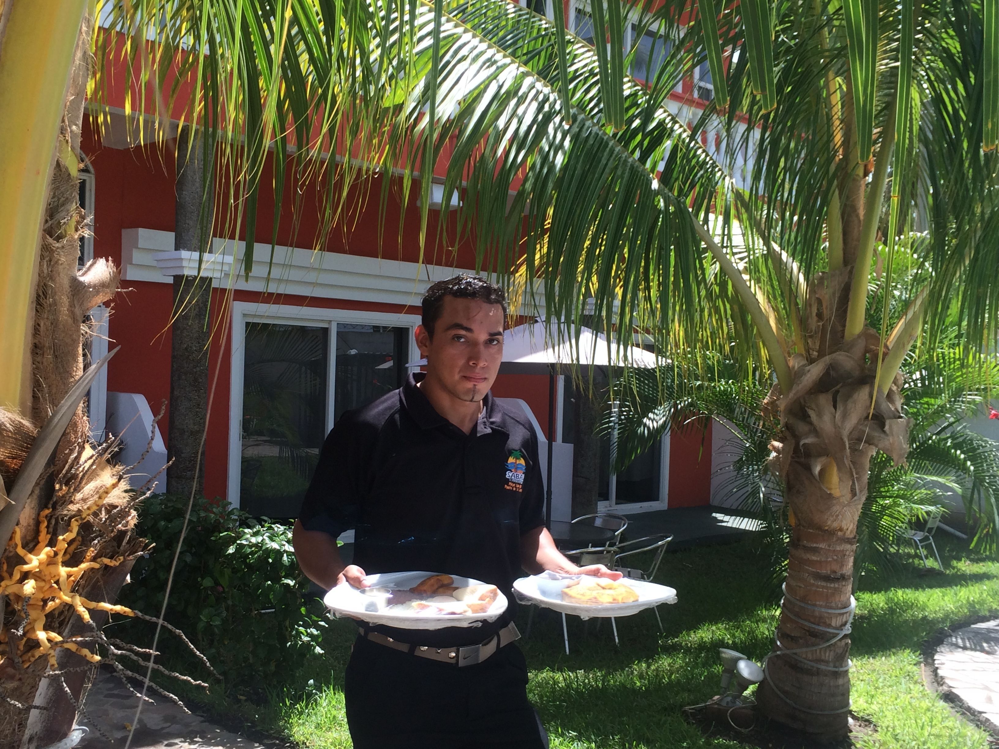 Una persona sostiene un plato con comida, cerca de una palmera y un edificio con fachada roja y blanca.