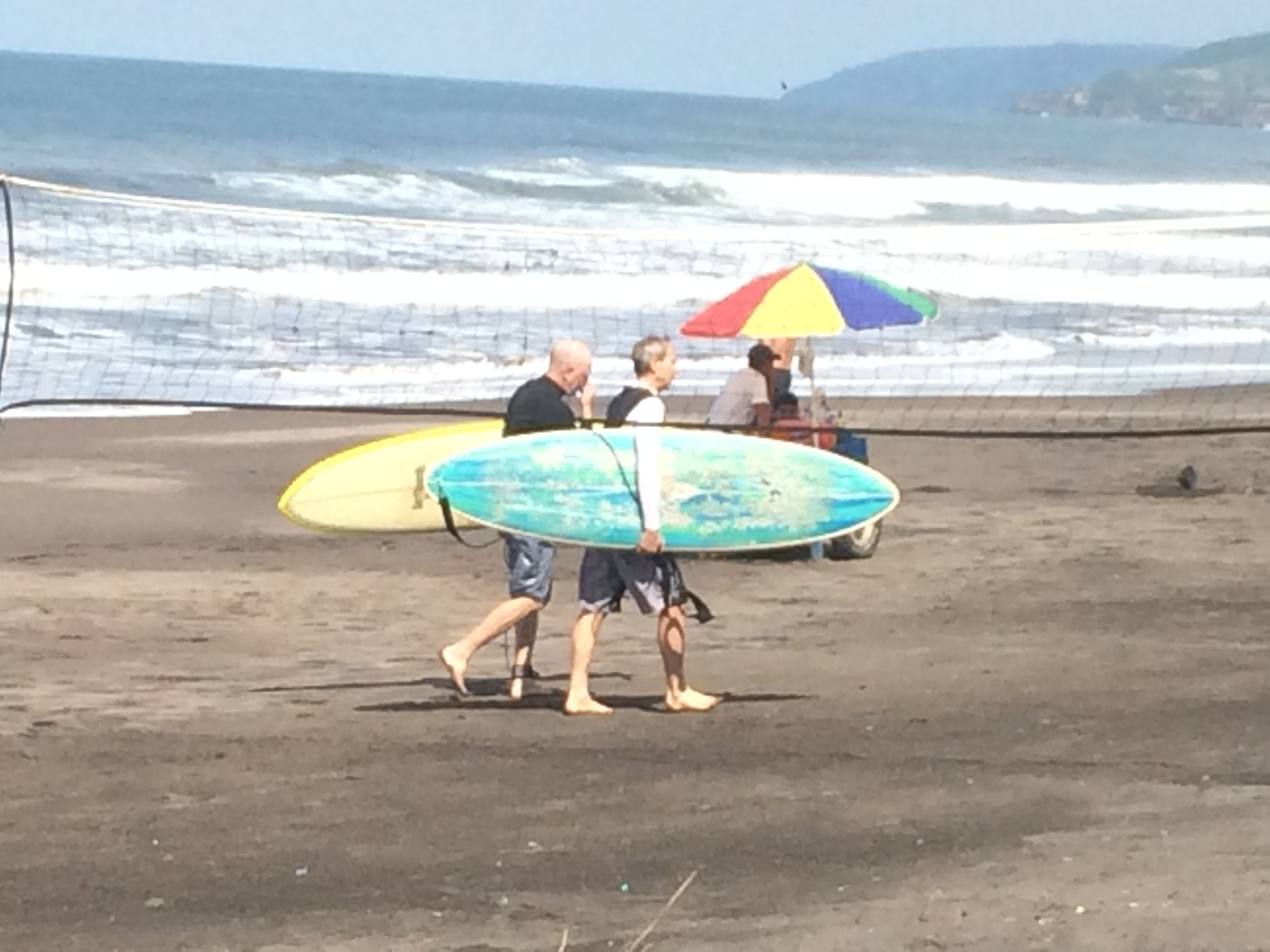 Dos personas caminando sobre una playa arenosa, cada una llevando una tabla de surf.