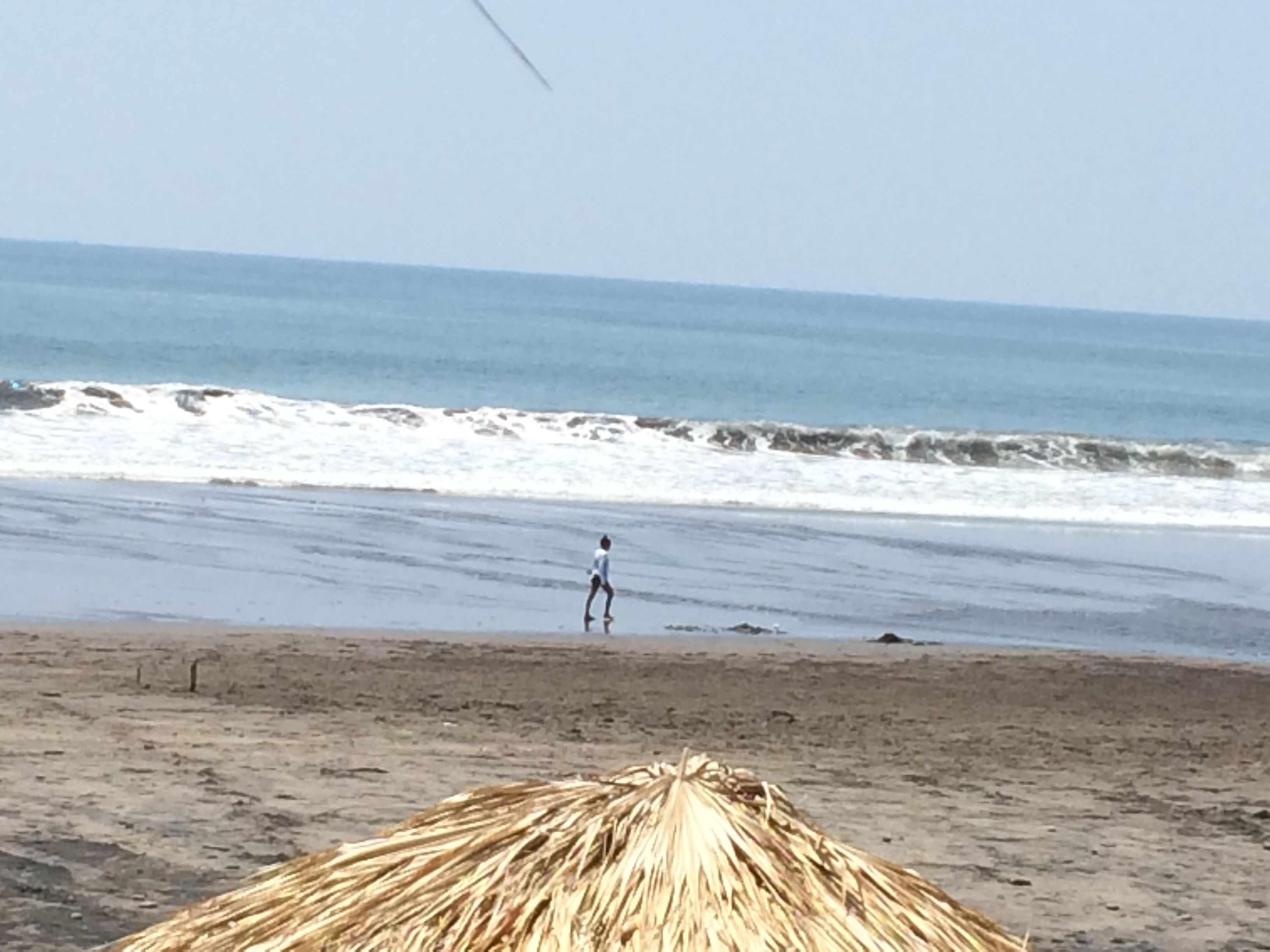 A beach with a person running towards the water, a thatched structure, and waves crashing onto the shore.