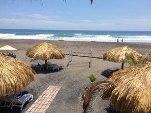 Una playa con sombrillas de paja, sillas de descanso y un camino empedrado.