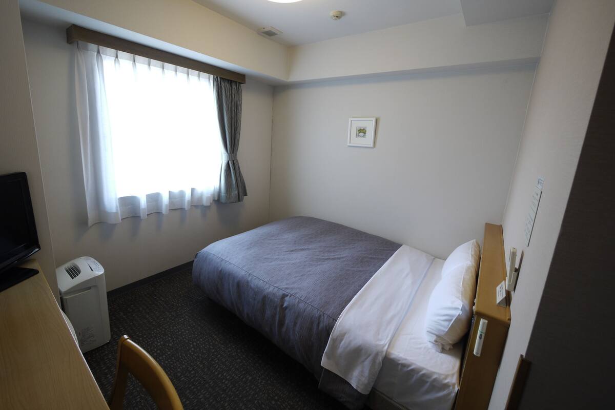 A hotel room with a single bed, a desk, a chair, a small heater, a window with curtains, and a wall-mounted light fixture.