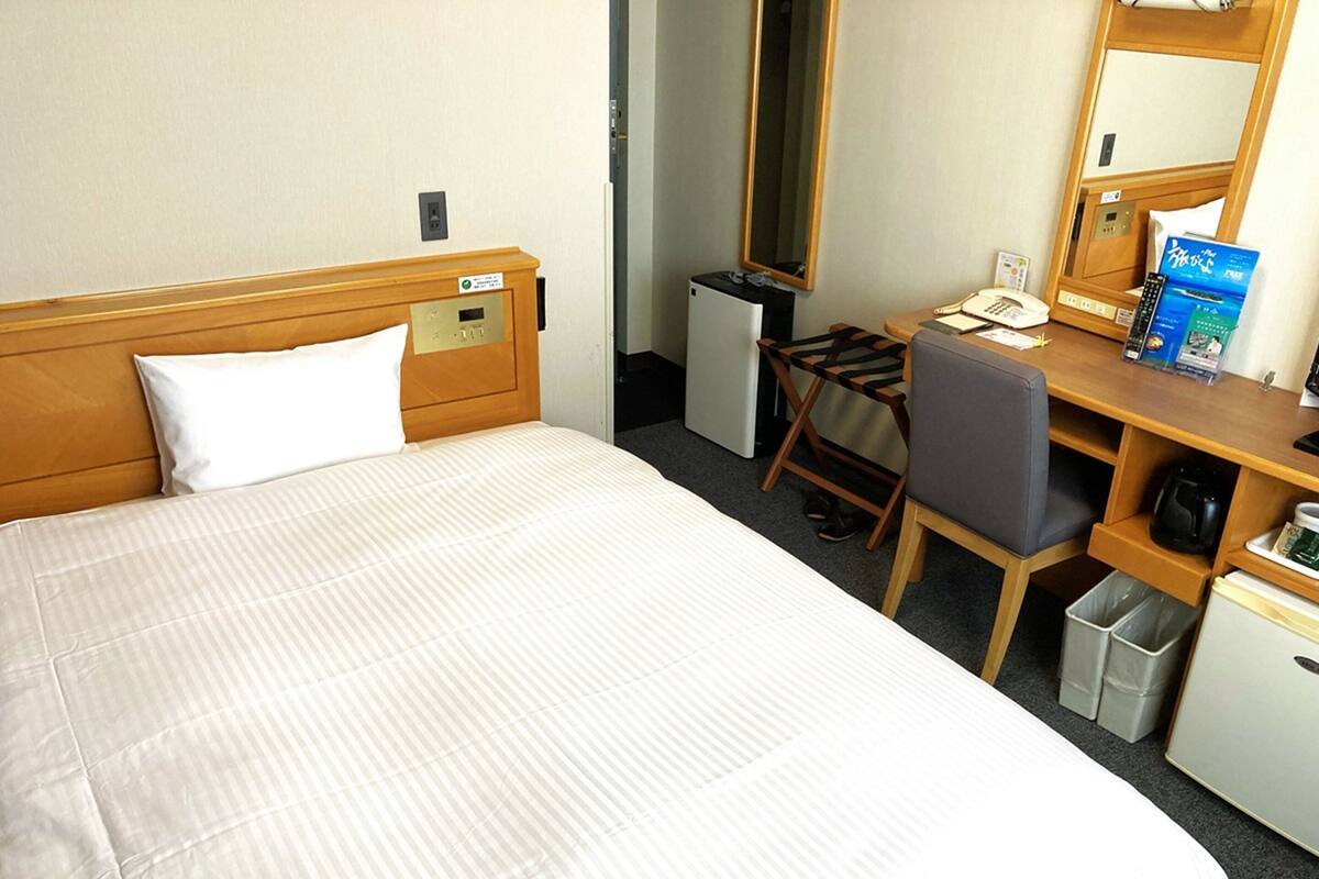 A hotel room with a bed, a desk, a chair, a small refrigerator, a trash bin, and a mirror.