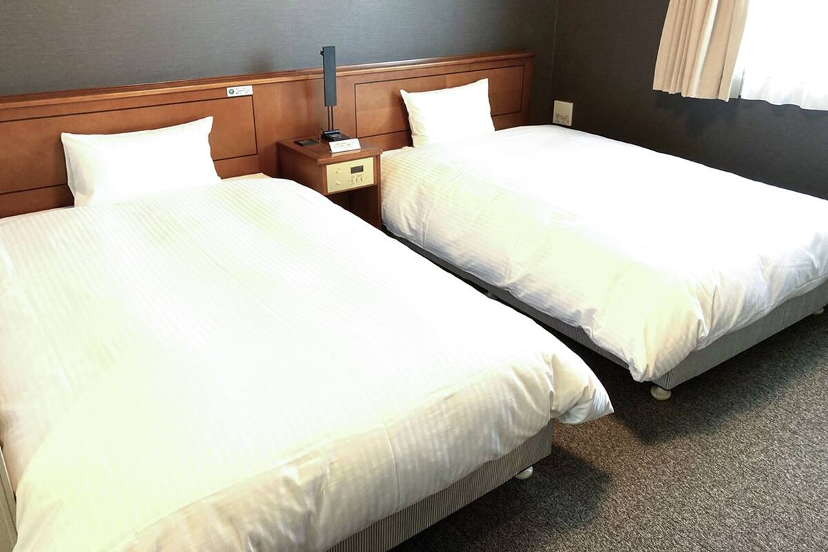 A hotel room with two single beds, a wooden headboard, a bedside table with a lamp, and a window with curtains.
