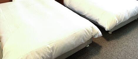 A hotel room with two single beds, a wooden headboard, a bedside table with a lamp, and a window with curtains.
