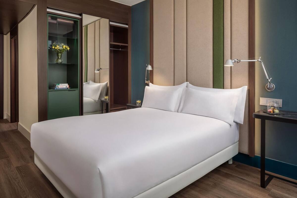 A modern hotel room with a large bed, a bedside table with a lamp, a mirror, and a green wall.
