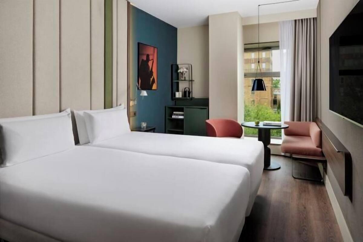 A hotel room with two beds, a desk, a chair, a TV, and a view of greenery.