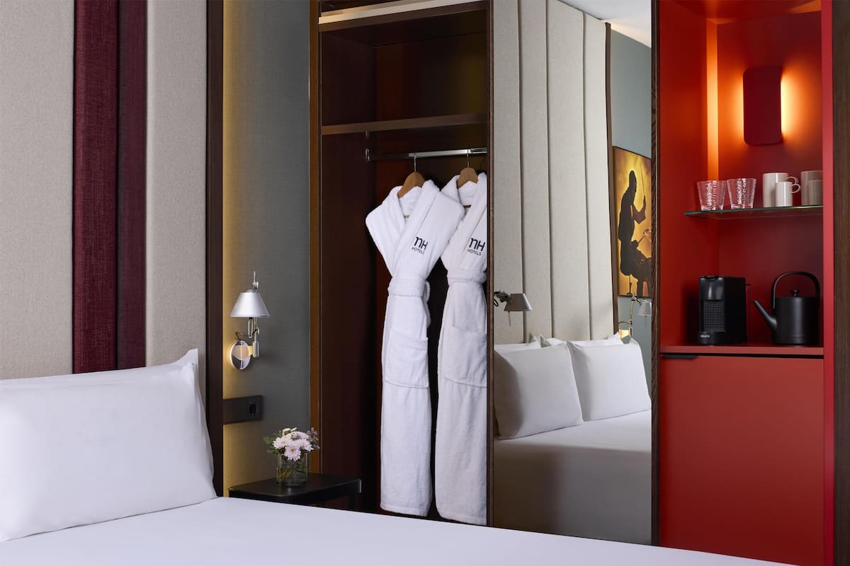 A hotel room with a bed, a nightstand with a lamp, a vase with flowers, a closet with robes, and a shelf with mugs and a kettle.