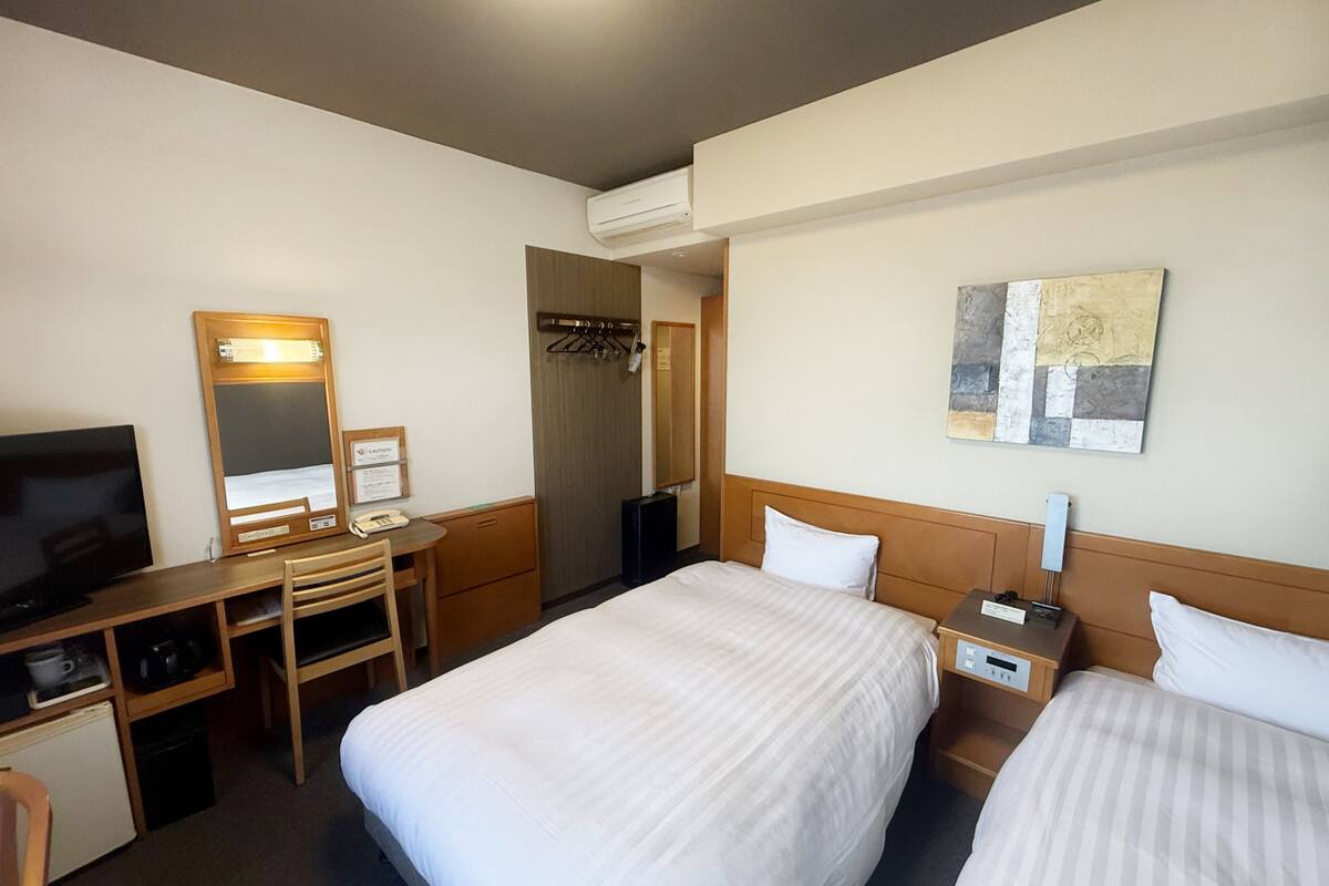 Une chambre d’hôtel avec deux lits, un bureau avec une chaise, une télévision, un porte-manteau et un tableau au mur.