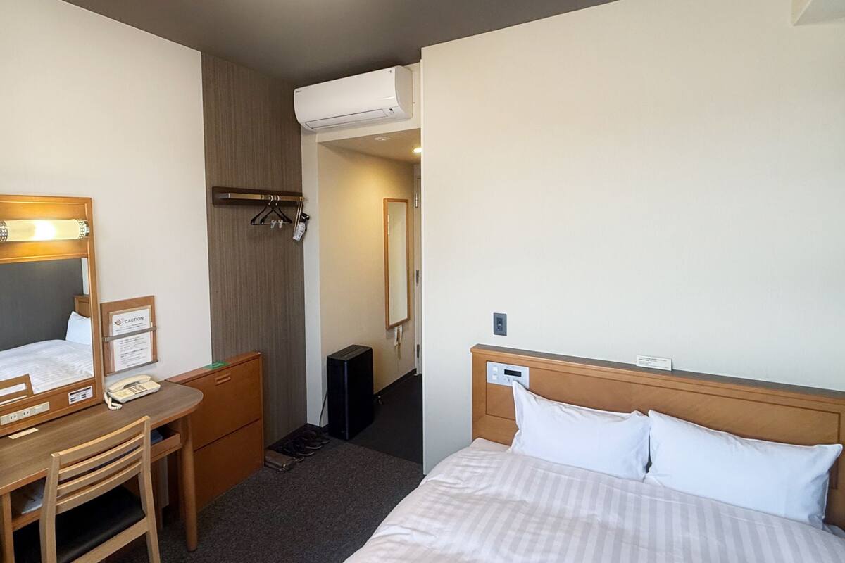Une chambre d’hôtel comprenant un lit, un bureau avec une chaise, un miroir, une armoire et un appareil de climatisation.