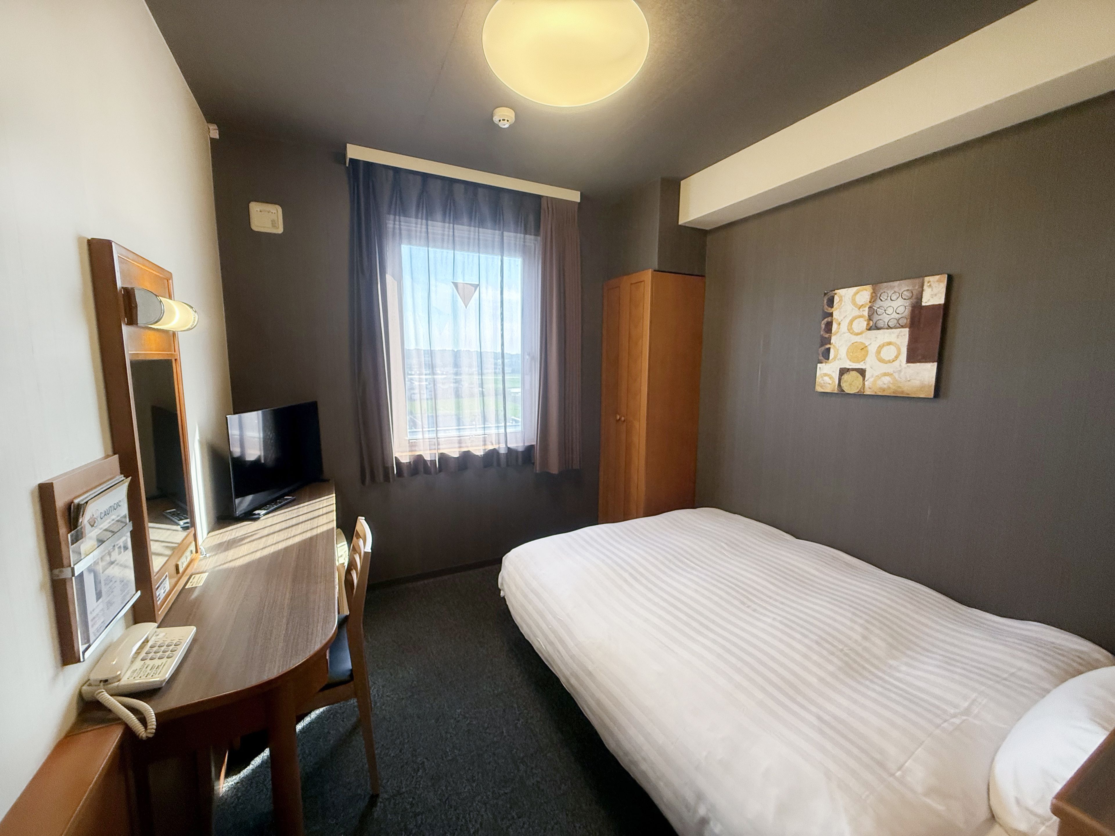 Una habitación de hotel con cama, escritorio con teléfono, televisor, ventana con cortinas y una obra de arte colgada en la pared.