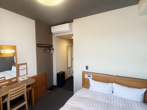 Ein Hotelzimmer mit Bett, Schreibtisch mit Stuhl, Spiegel und Kleiderschrank sowie einer Klimaanlage.