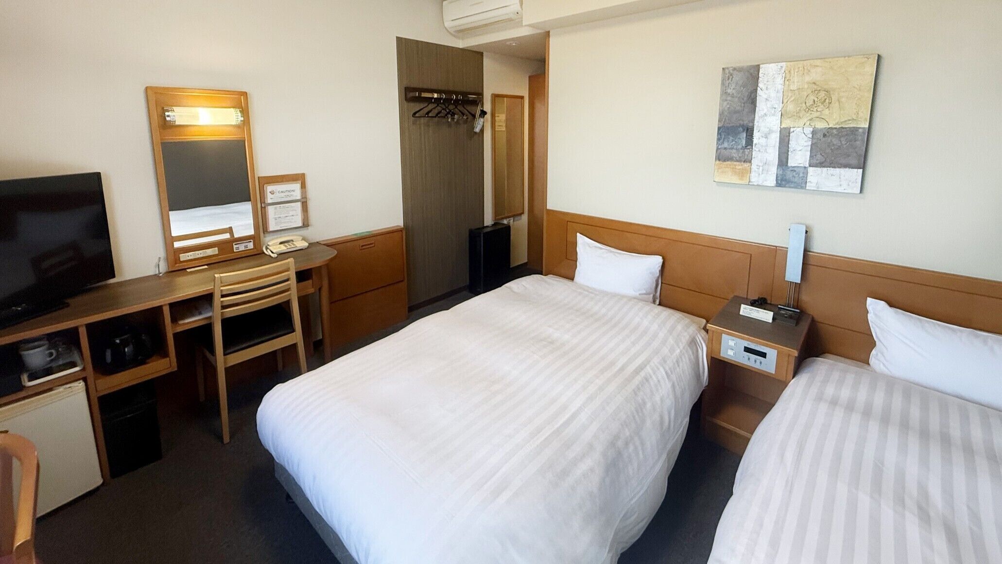 Ein Hotelzimmer mit zwei Betten, einem Schreibtisch mit Stuhl, einem Fernseher, einem Spiegel und einem Wandbild.
