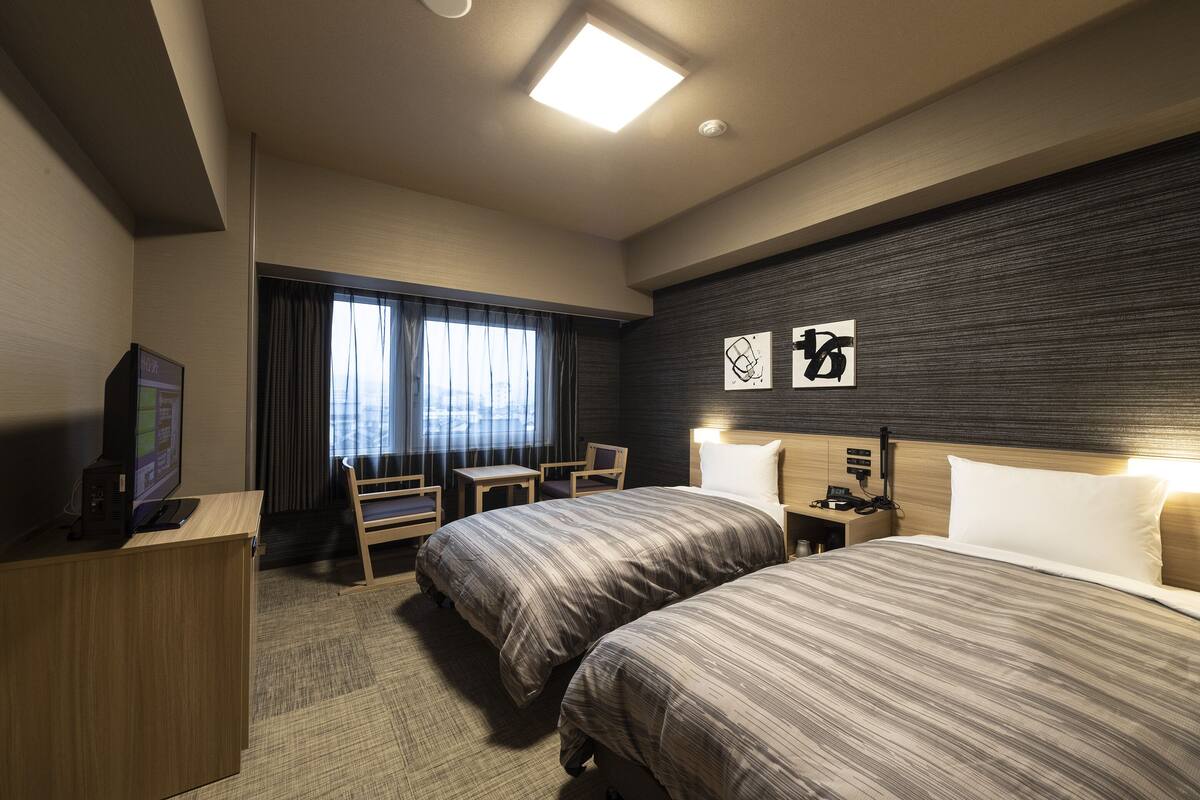 A hotel room with two beds, a desk with a computer, a TV, and a window with curtains.