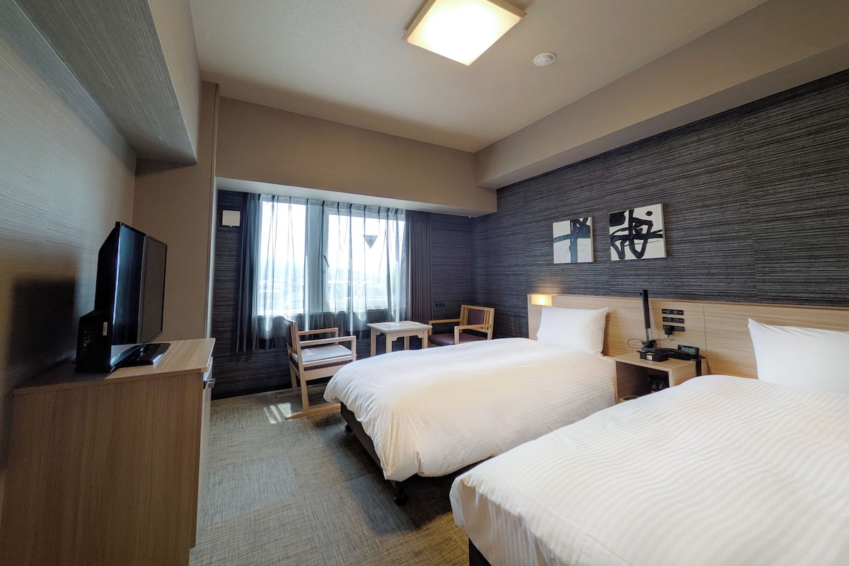 A hotel room with two beds, a desk, a chair, a television, and artwork on the wall.