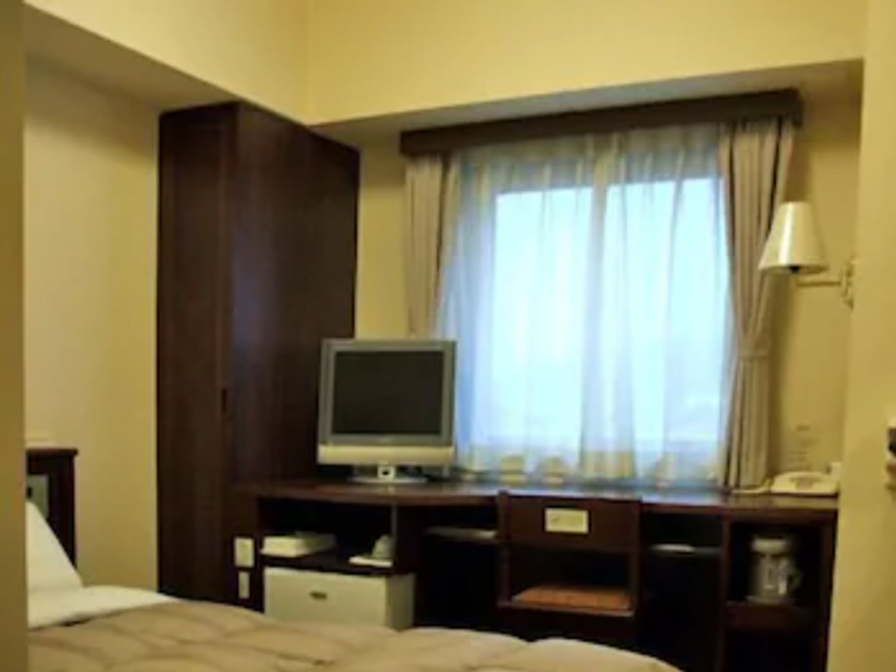 A hotel room with a bed, a desk with a computer, a chair, a window with curtains, and a lamp.