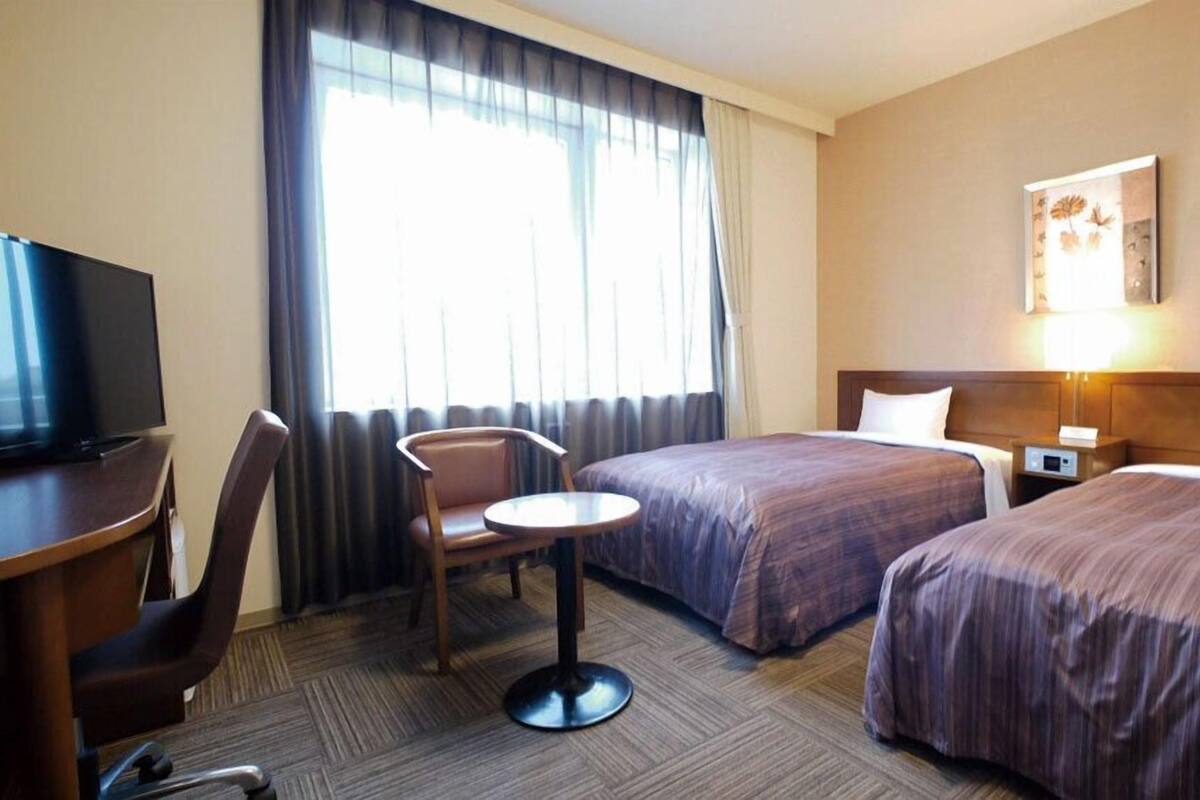 Ein Hotelzimmer mit zwei Betten, einem Schreibtisch mit Fernseher, einem Sessel, einem kleinen Tisch und einem Fenster mit Vorhängen.