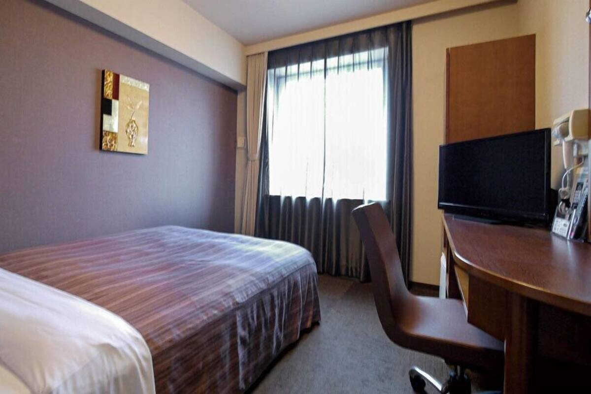 Ein Hotelzimmer mit einem Bett, einem Schreibtisch mit Stuhl, einem Fernseher und einem Fenster mit Vorhängen.