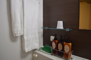 A bathroom with a white towel hanging on the wall, a glass shelf with a white cup, and three bottles of body soap and shampoo. - Hotel Route Inn Yatsushiro (Yatsushiro)